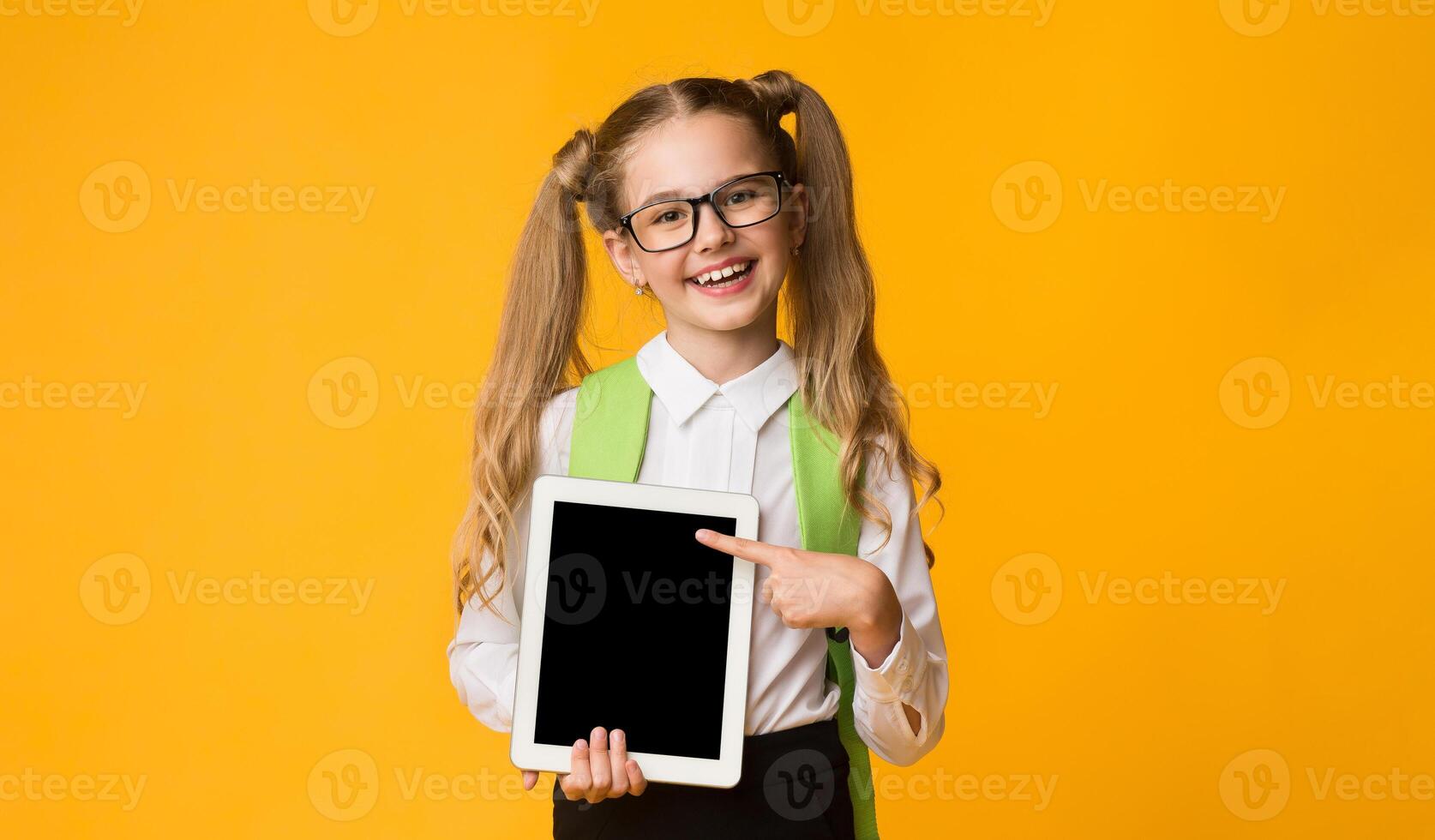 E-Learning. Elementary Student Girl With Digital Tablet Showing Blank Computer Screen Posing On Yellow Studio Background. Mockup photo