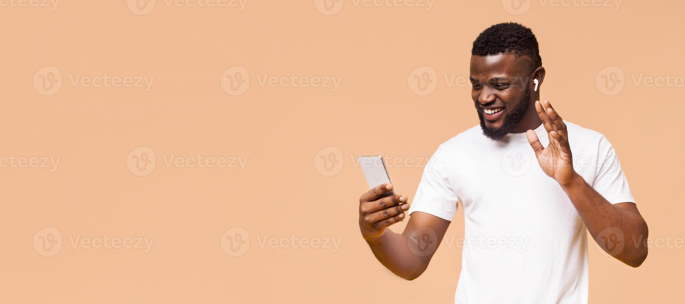 A young African American man in a white t-shirt smiles and waves as he uses his smartphone for a call. The background is a simple, soft beige, copy space photo