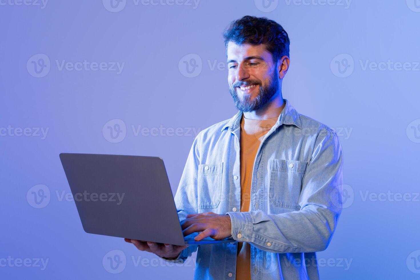 A man wearing a blue shirt is sitting and actively using a laptop computer. His hands are positioned on the keyboard, and his gaze is focused on the screen as he works. photo