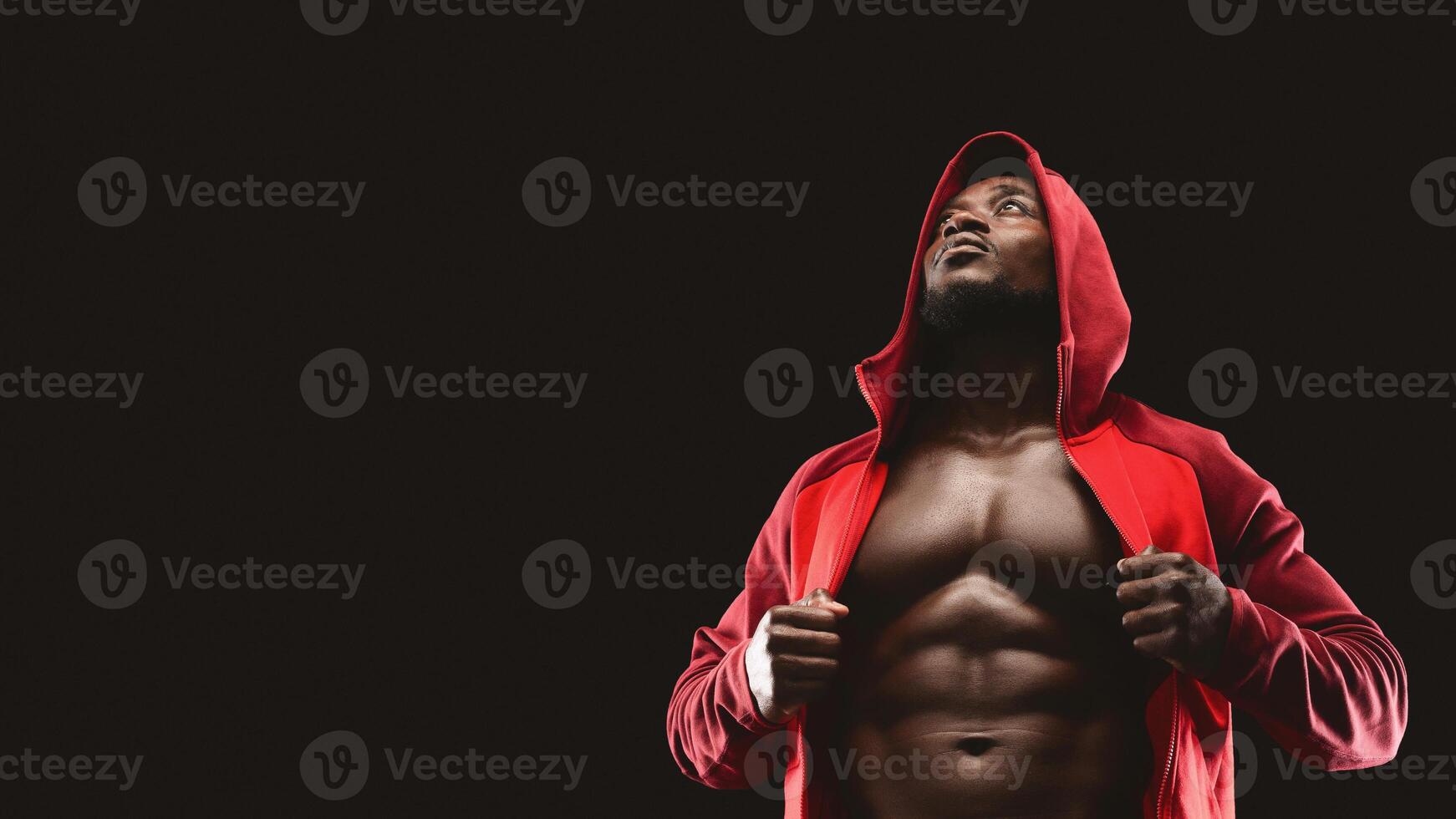 A muscular man in a red hoodie stands against a black background. He is looking up and revealing his toned chest. photo
