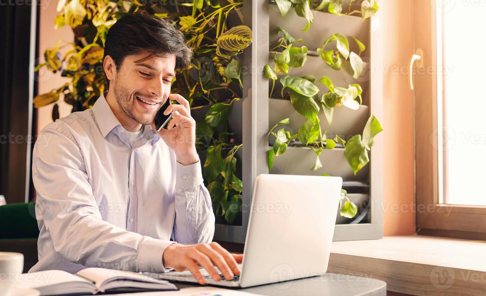 Small business development. Cheerful businessman working on laptop and talking on phone in cafe, free space photo
