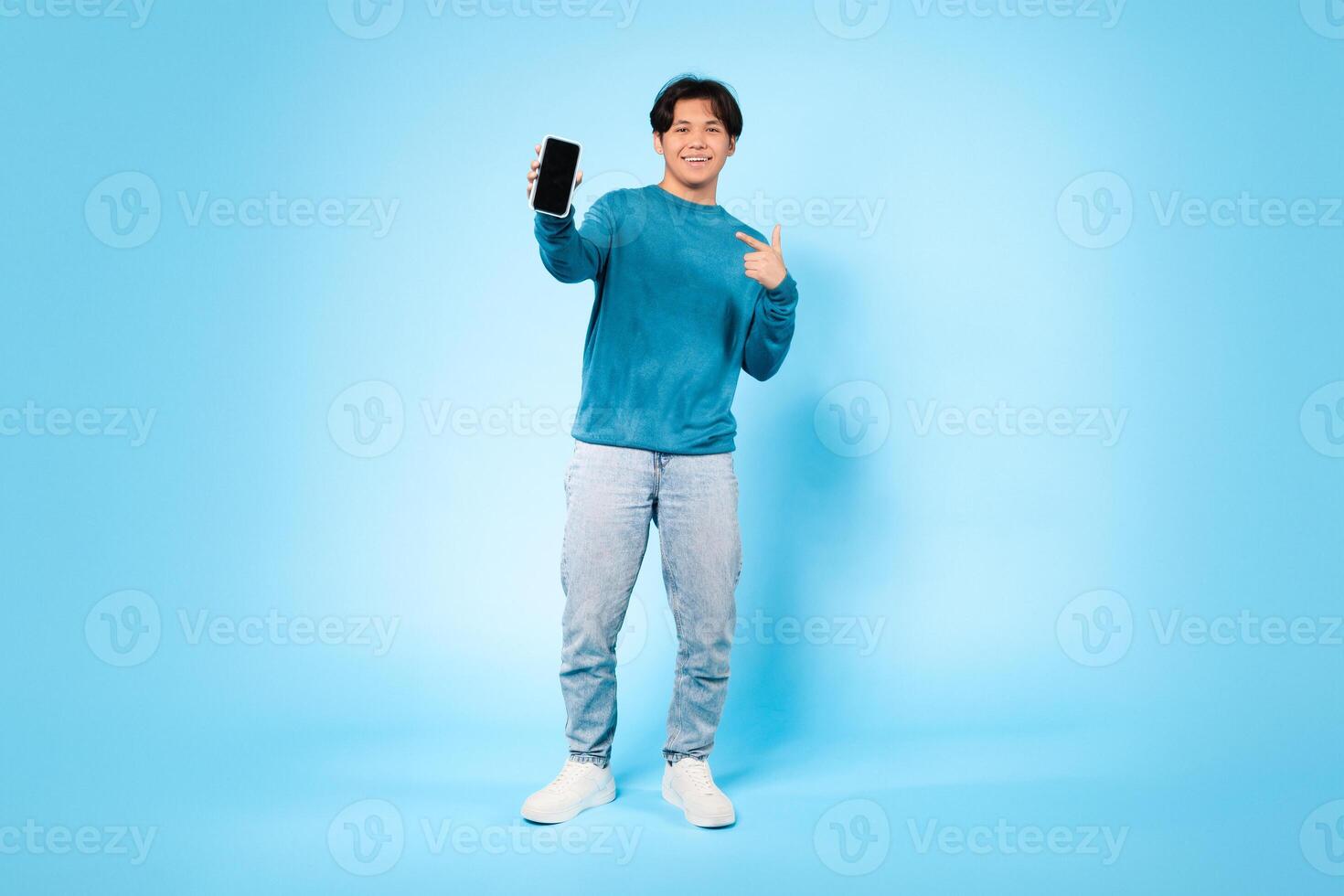 Asian young guy is standing in a brightly lit studio, smiling and pointing to a smartphone he is holding in his left hand. The vibrant blue background photo