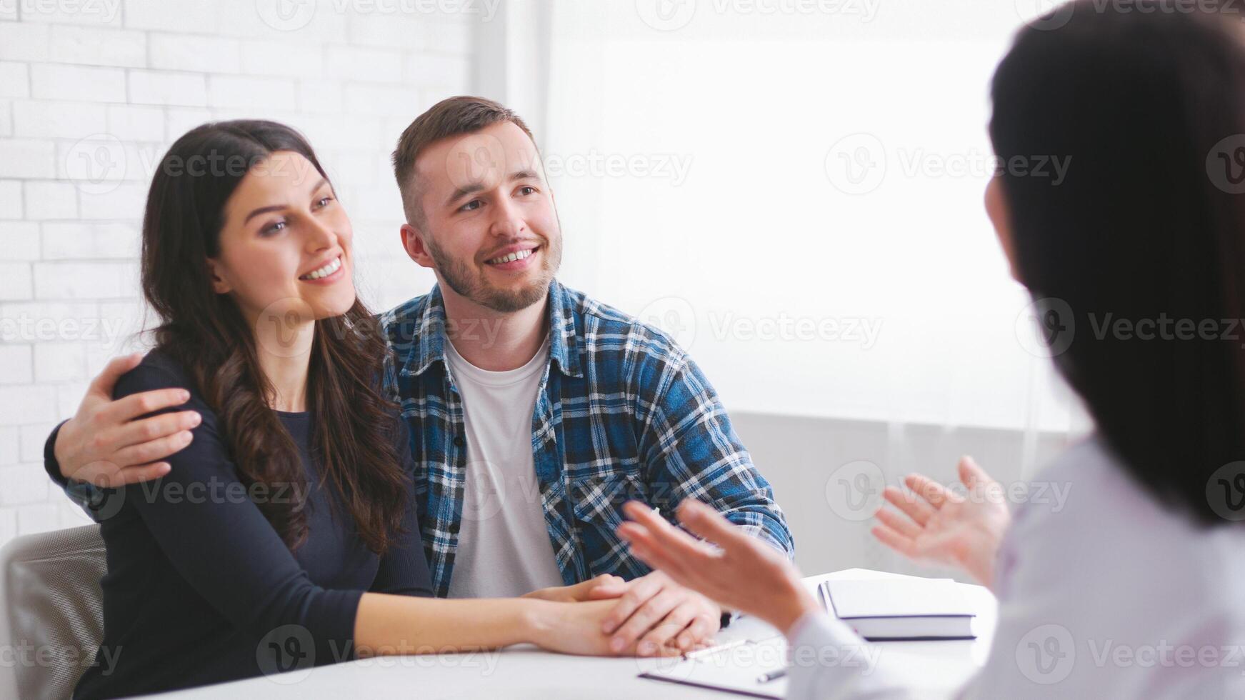 A young couple sits at a table listening attentively as a financial advisor explains something to them. The couple is smiling and appears to be engaged in the conversation photo