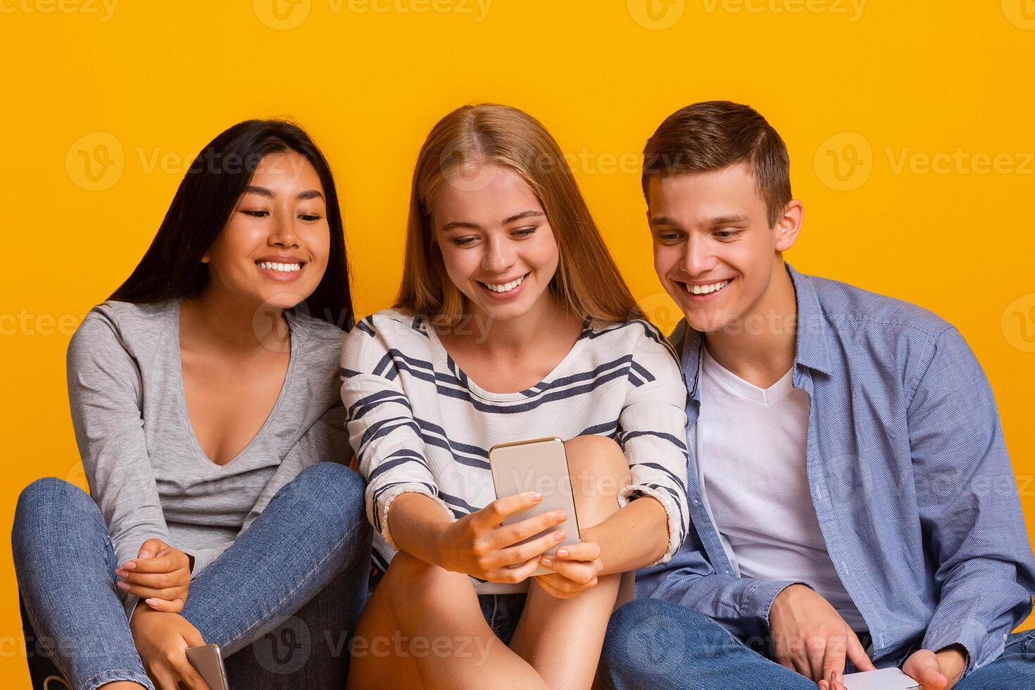 Group of multiracial classmates having break, watching videos on smartphone over yellow background photo