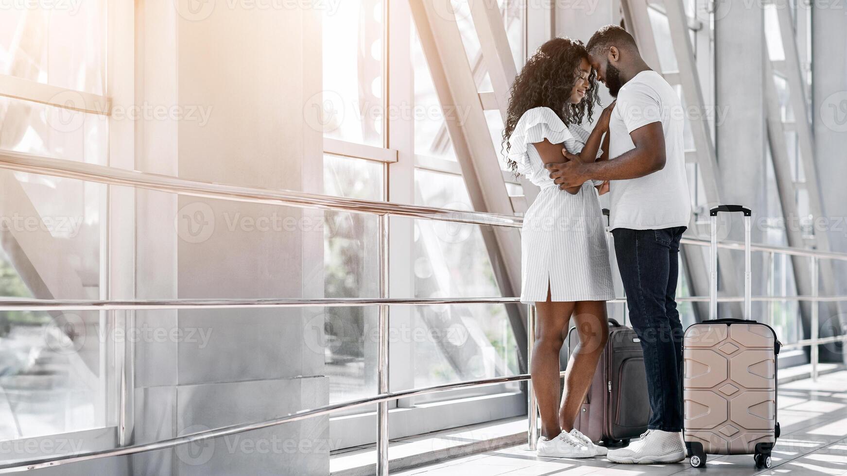 A young black couple embraces in an airport departure area, the man with his arm around the womans waist, their foreheads touching. They appear to be saying goodbye, with luggage visible behind them. photo
