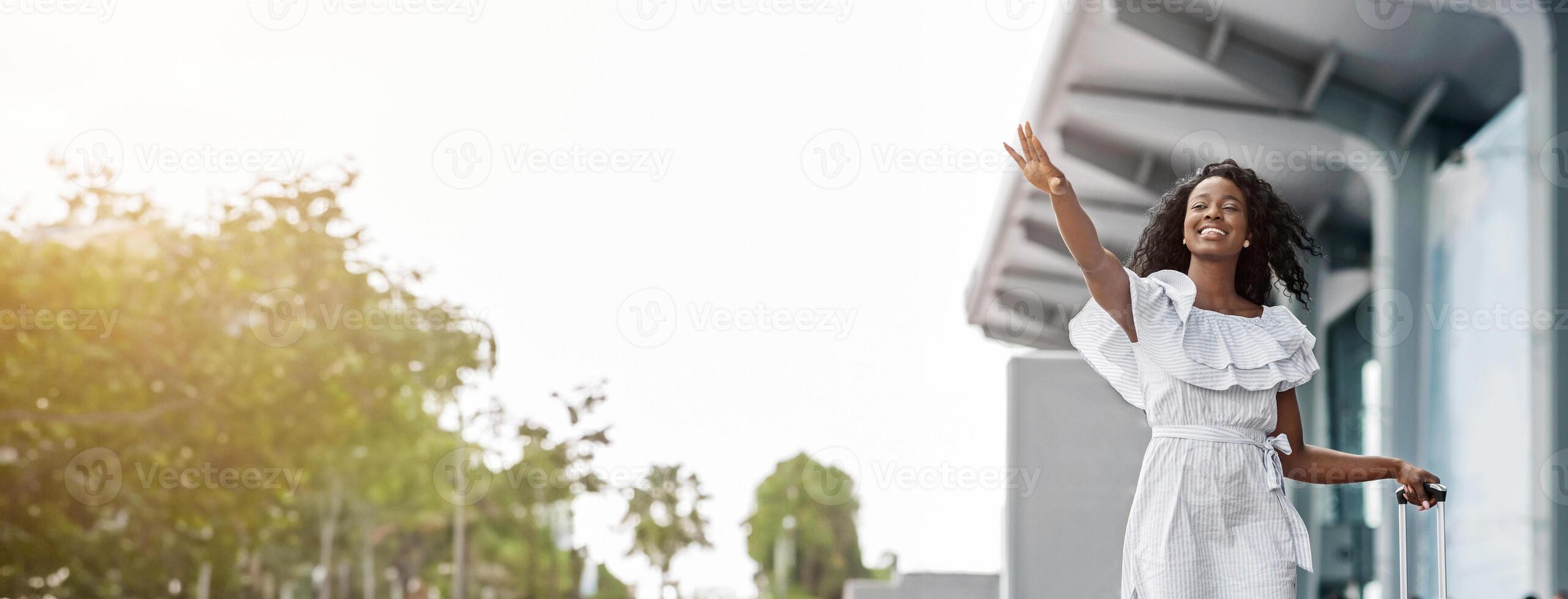 A happy black woman wearing a white dress, waves goodbye while holding a suitcase at an airport. She is standing outside, under a modern awning and the sun is shining, copy space photo