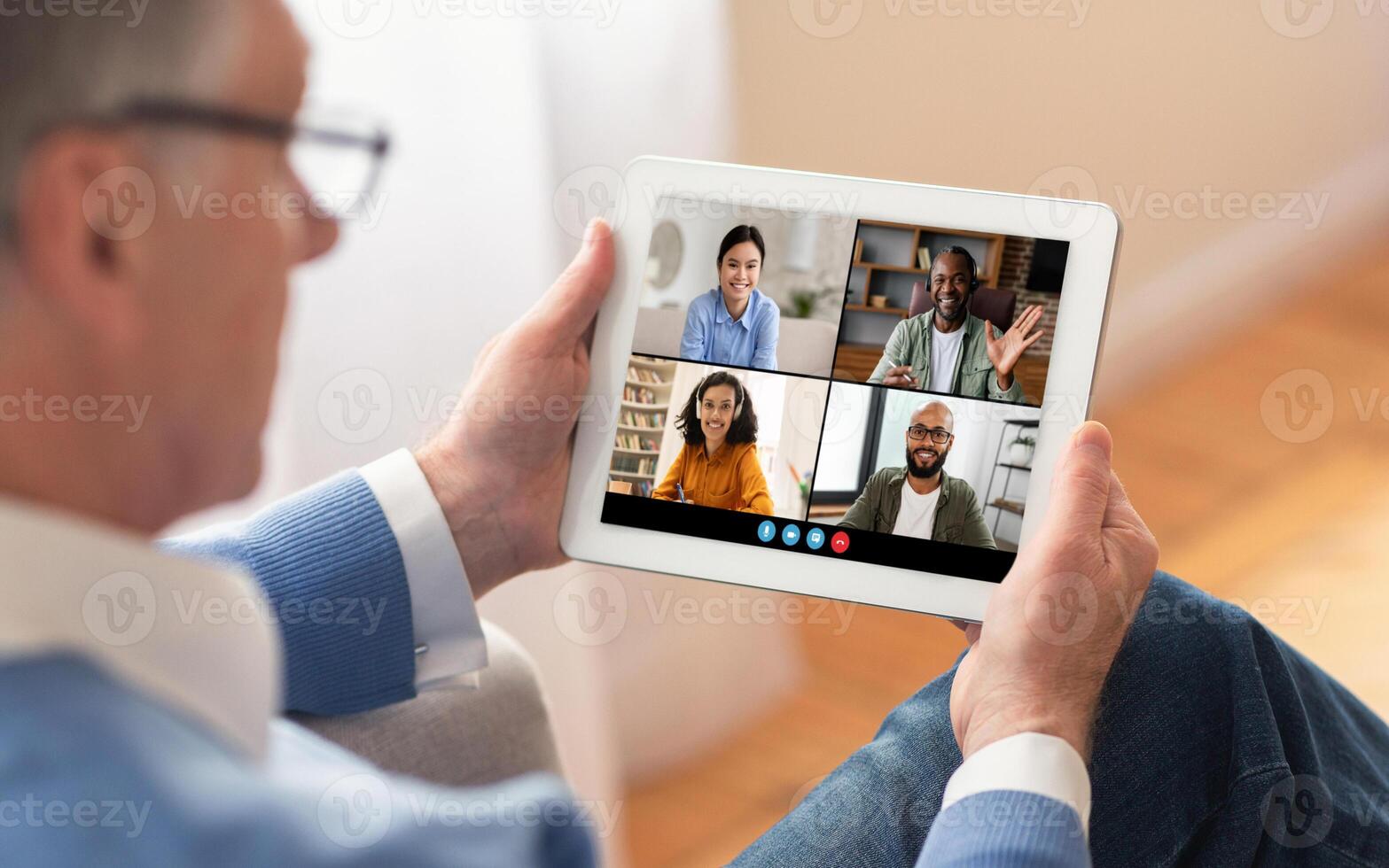 An older man wearing eyeglasses is sitting on a couch, holding a tablet and participating in a conference with three other colleagues photo