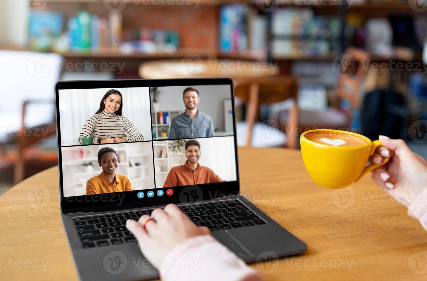 A person sits in a cafe, using a laptop to conference with three colleagues. They are smiling and engaged in conversation, enjoying a cup of coffee. The cafe is cozy and relaxed photo