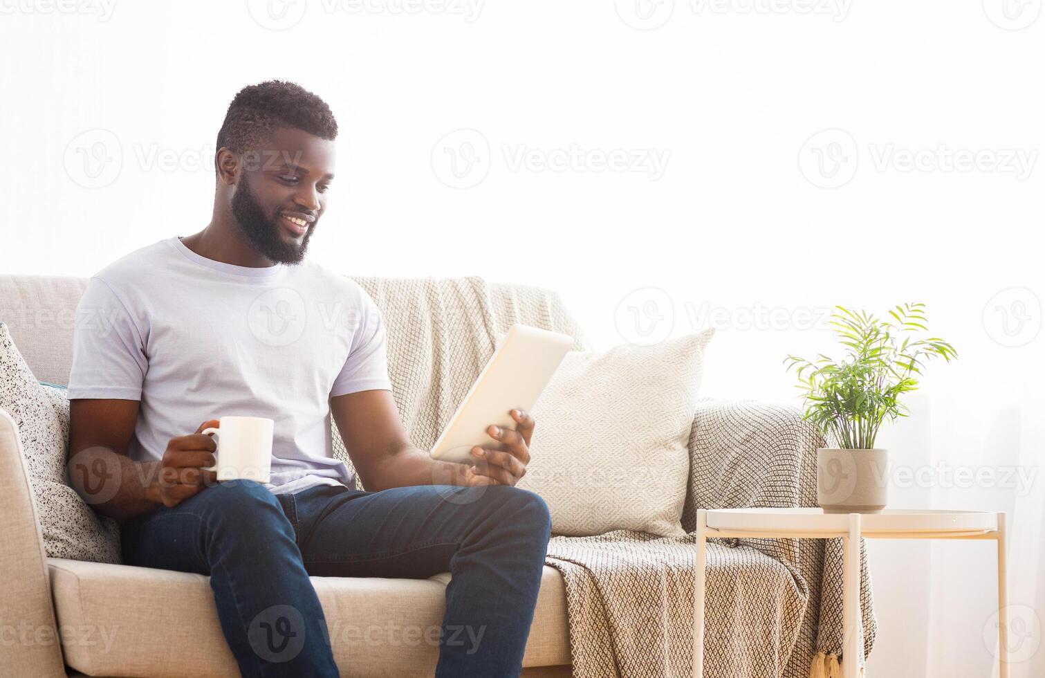 A young black man with a beard sits on a beige sofa in a bright living room, holding a white tablet and a cup of coffee in his hands. He is looking down at the tablet, smiling photo