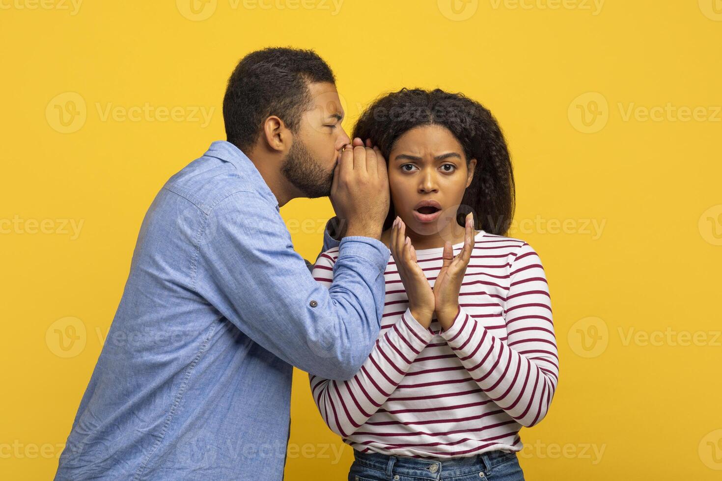African American man dressed in a blue shirt is whispering into the ear of a young woman who looks surprised and shocked, isolated on yellow background photo