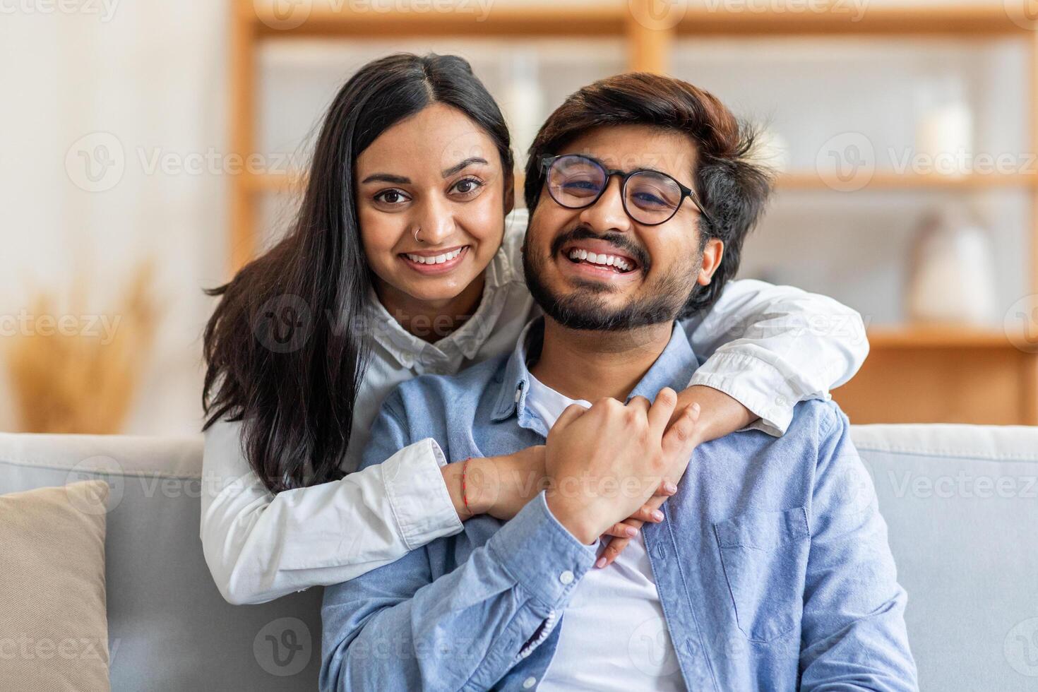 A joyful Indian couple embracing and smiling while sitting on a comfortable sofa in a warmly lit living room. They enjoy a moment of togetherness, creating a heartwarming, relaxed atmosphere. photo