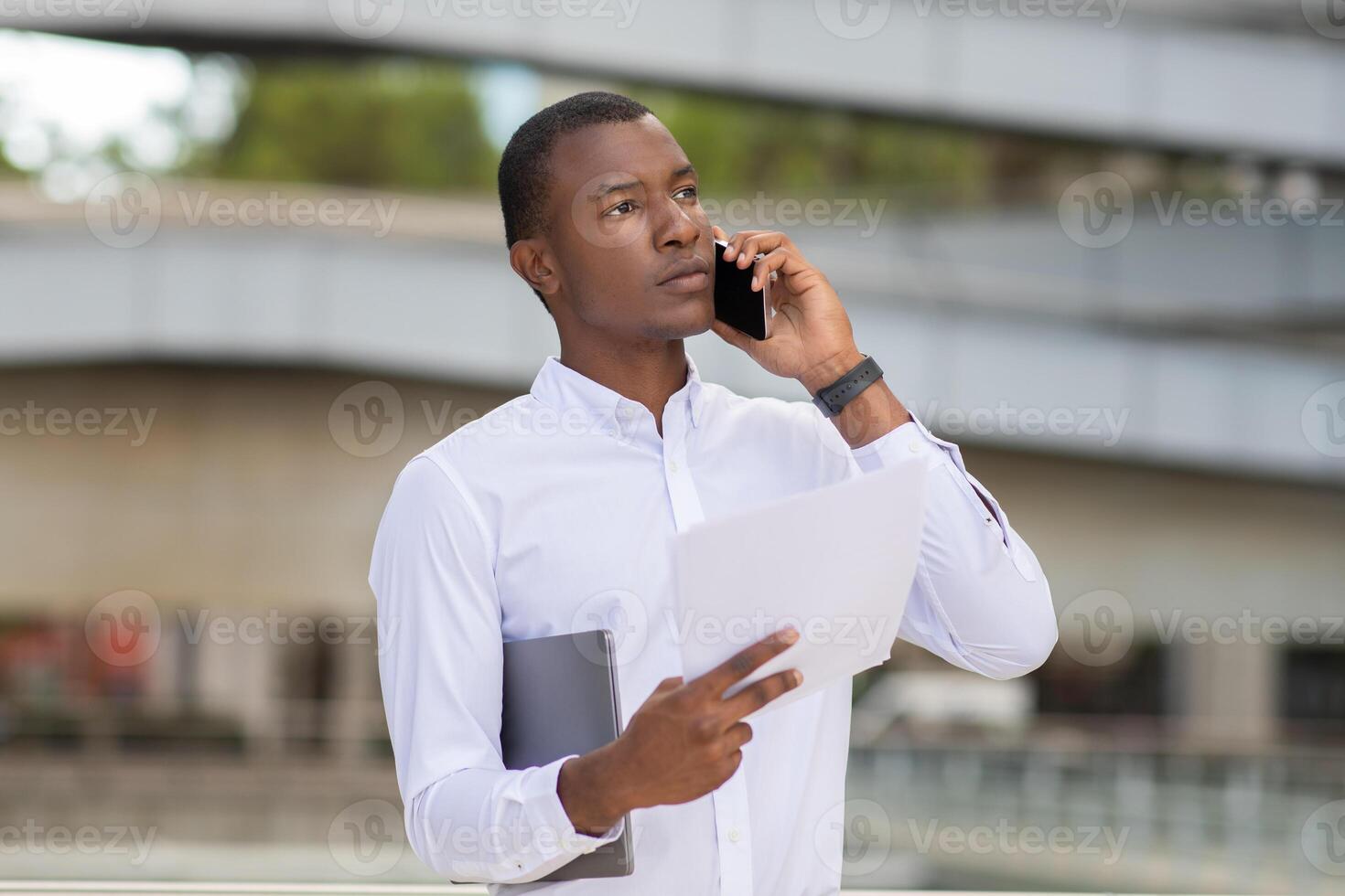 A young African American man stands in an urban environment, engaged in a phone conversation. He appears focused as he holds several documents in one hand and a tablet in the other. photo