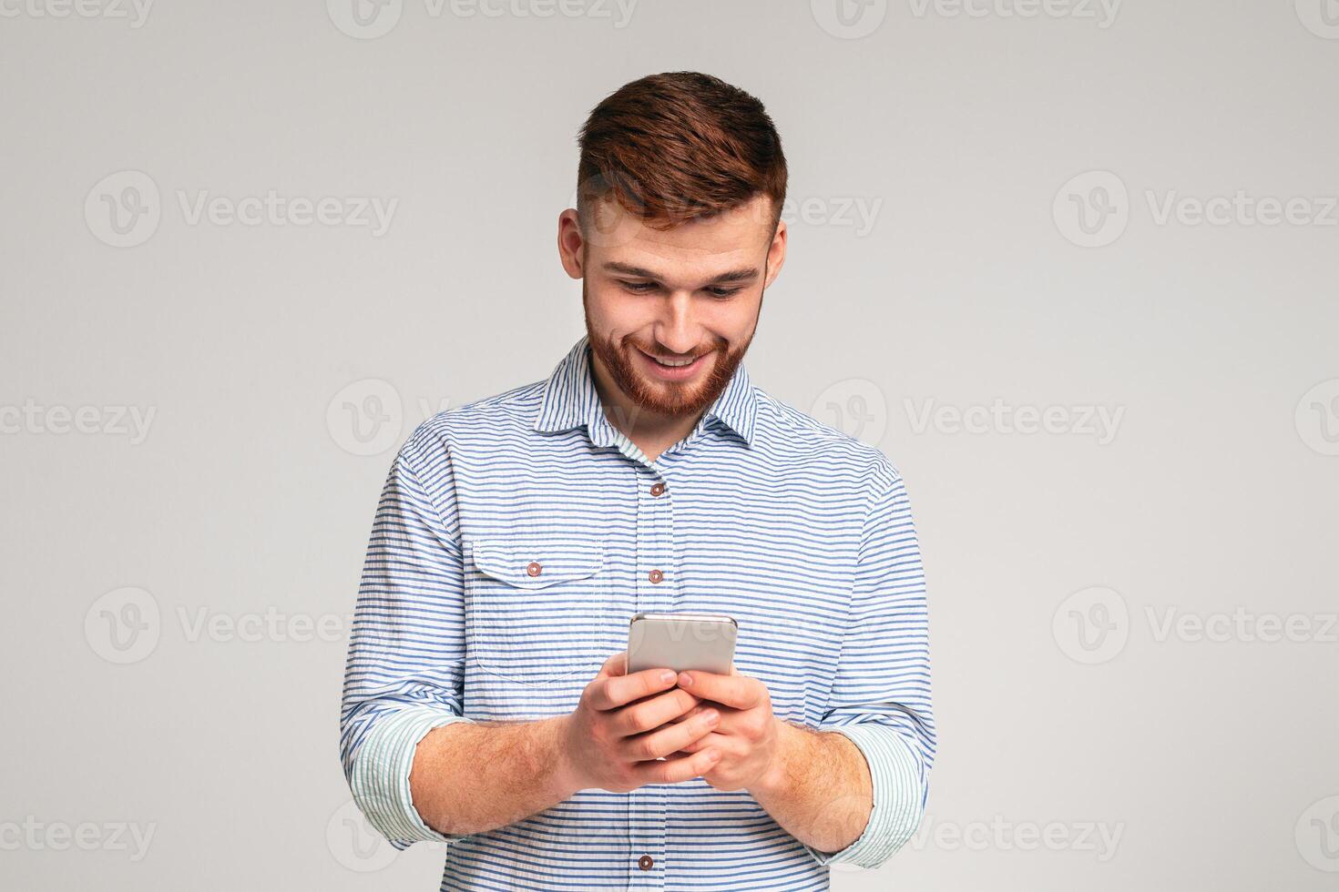 Young man absorbedly text messaging on his cellphone, isolated on grey studio background, free space photo