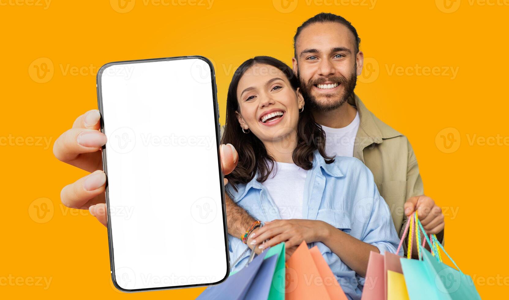 A smiling couple stands against a vibrant yellow background. They are both holding shopping bags and a smartphone with a blank screen. photo
