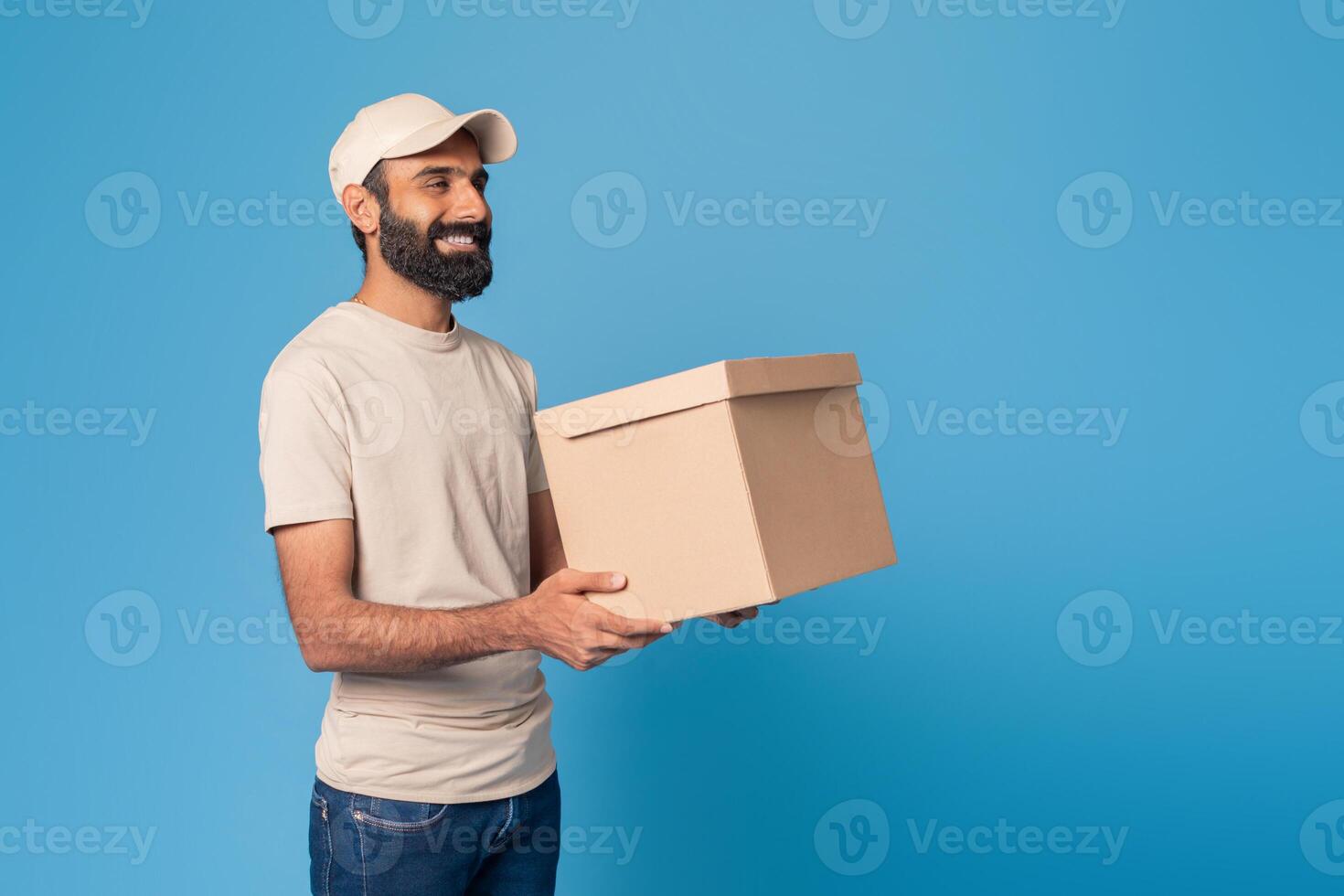 A smiling bearded Indian delivery man in a light cap and beige t-shirt holds a brown box in front of a bright blue background, copy space photo