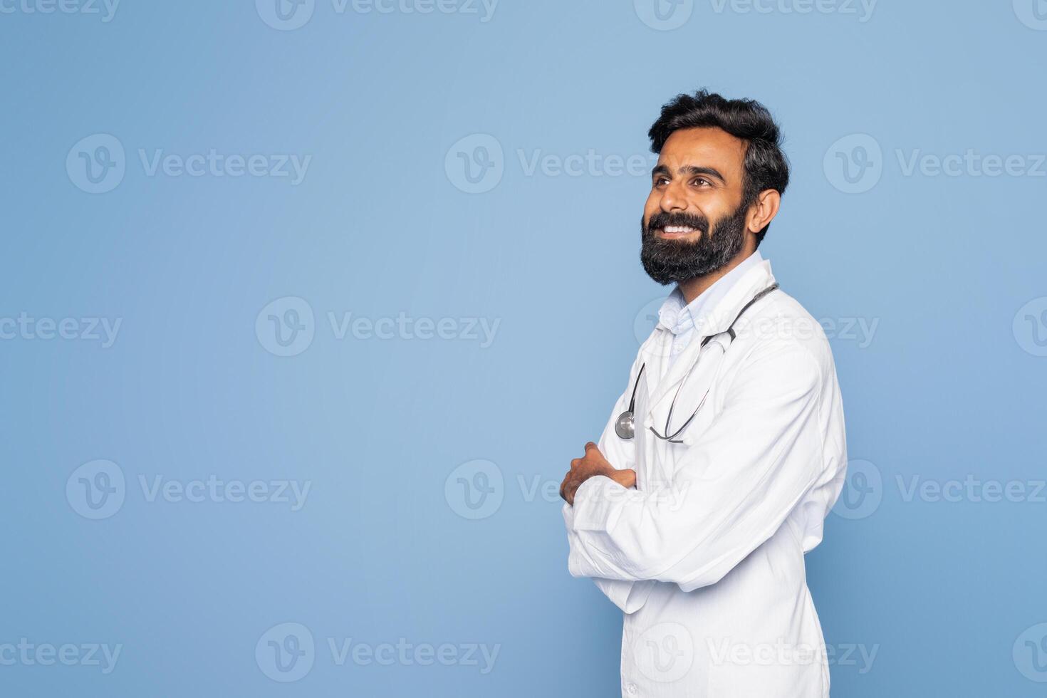 A bearded Indian doctor wearing a white coat and stethoscope smiles while standing confidently against a blue studio background. His arms are crossed, conveying professionalism and warmth. photo