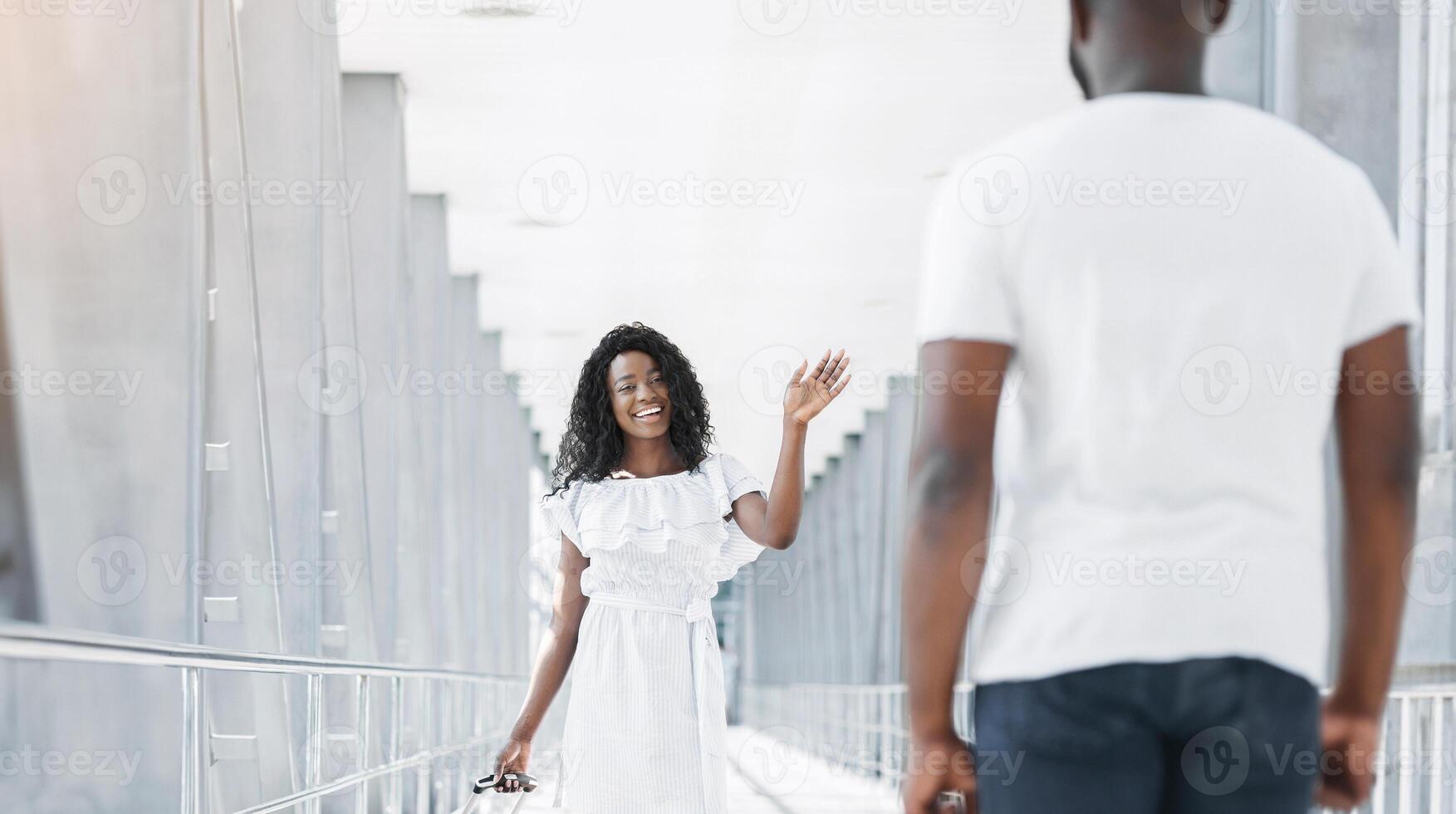 Black woman in a white dress waves goodbye to a man in a white shirt in an airport terminal. The man is turned away from the camera, and the woman is looking directly at the camera with a smile. photo