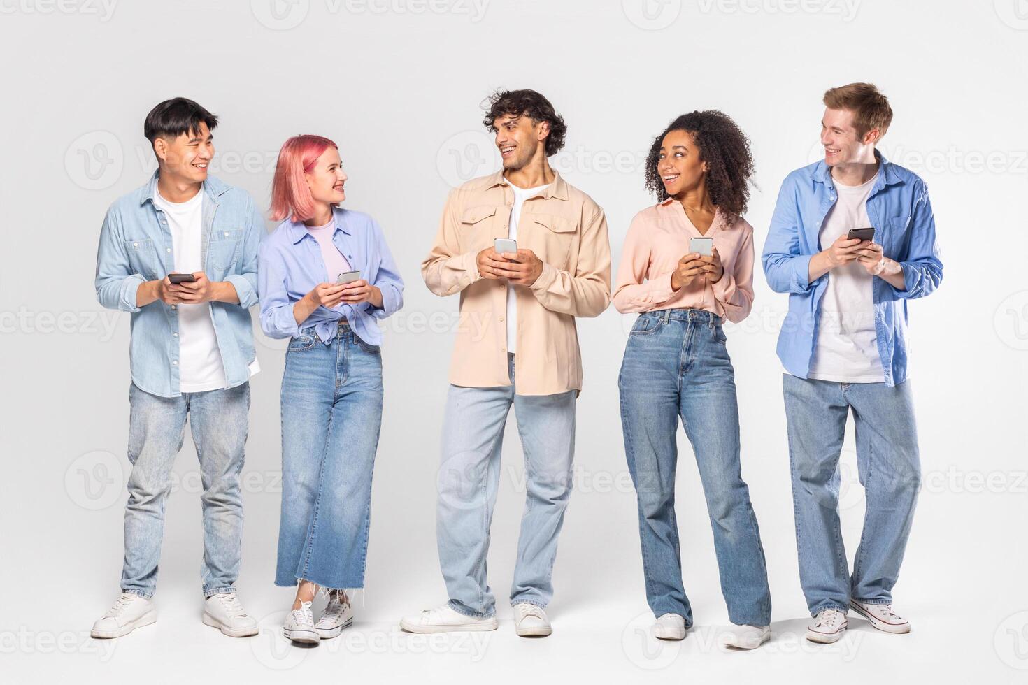 A group of five young adults, each wearing casual clothing and holding smartphones, stand in a line against a white background. They are all smiling and appear to be engaged in conversation. photo