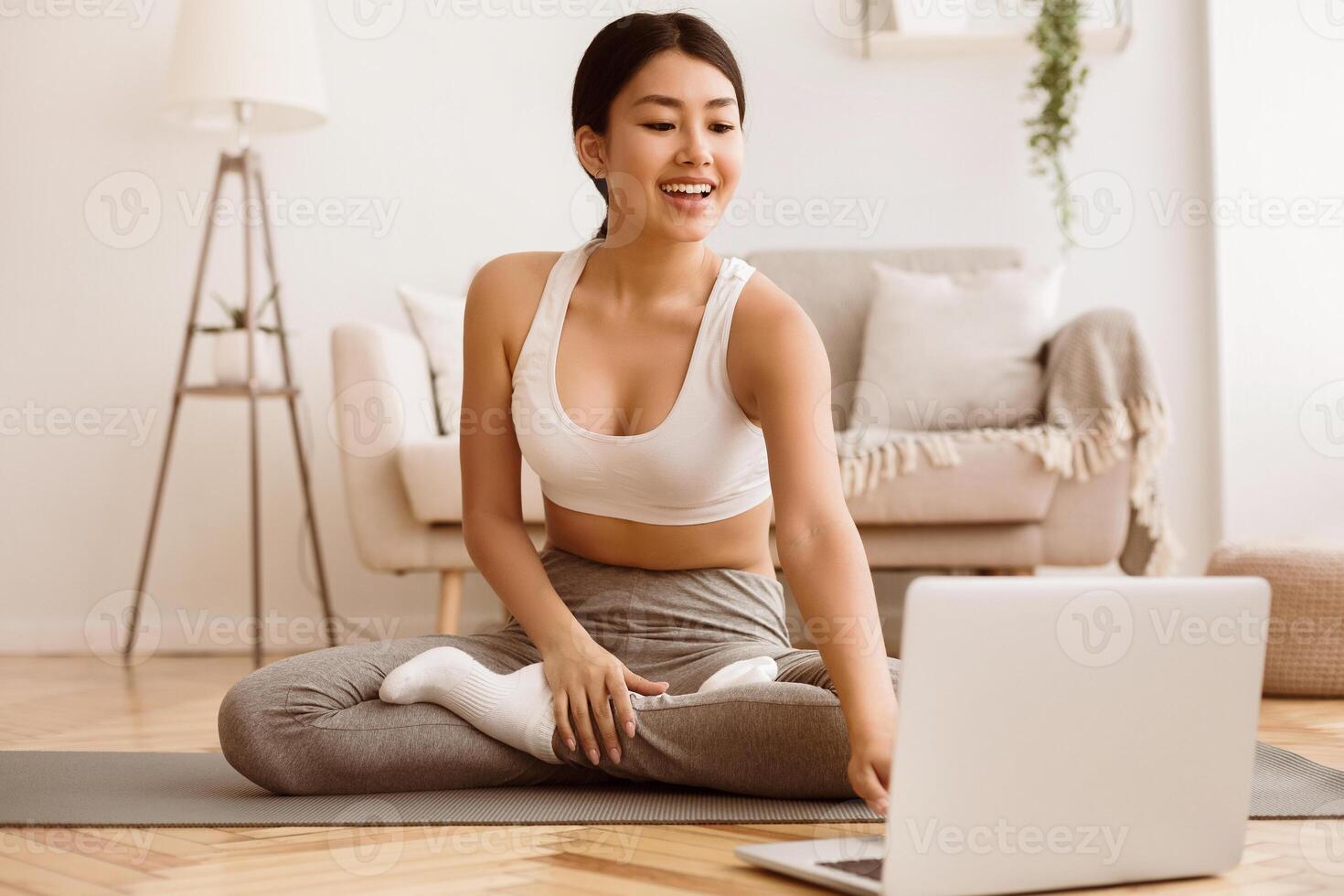 A young woman in a white sports bra and grey pants is sitting on a yoga mat in a living room, following along with a yoga on her laptop. photo