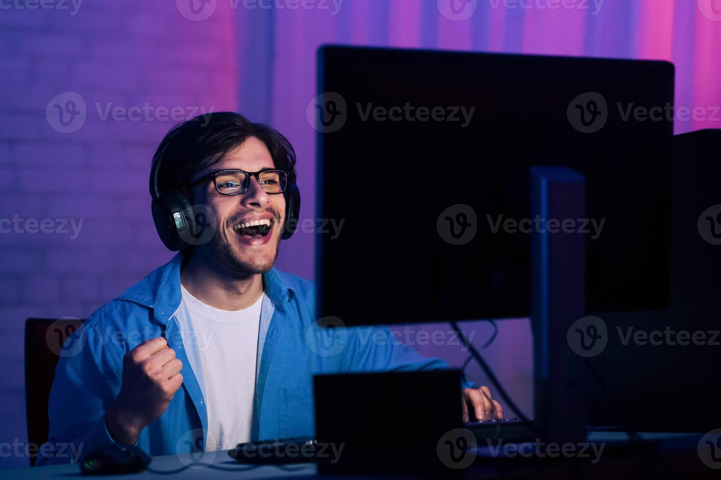 A young man, wearing glasses and headphones, is sitting in front of a computer screen in a dimly lit room. He is smiling and laughing, with his fist raised in the air, as he plays a game. photo