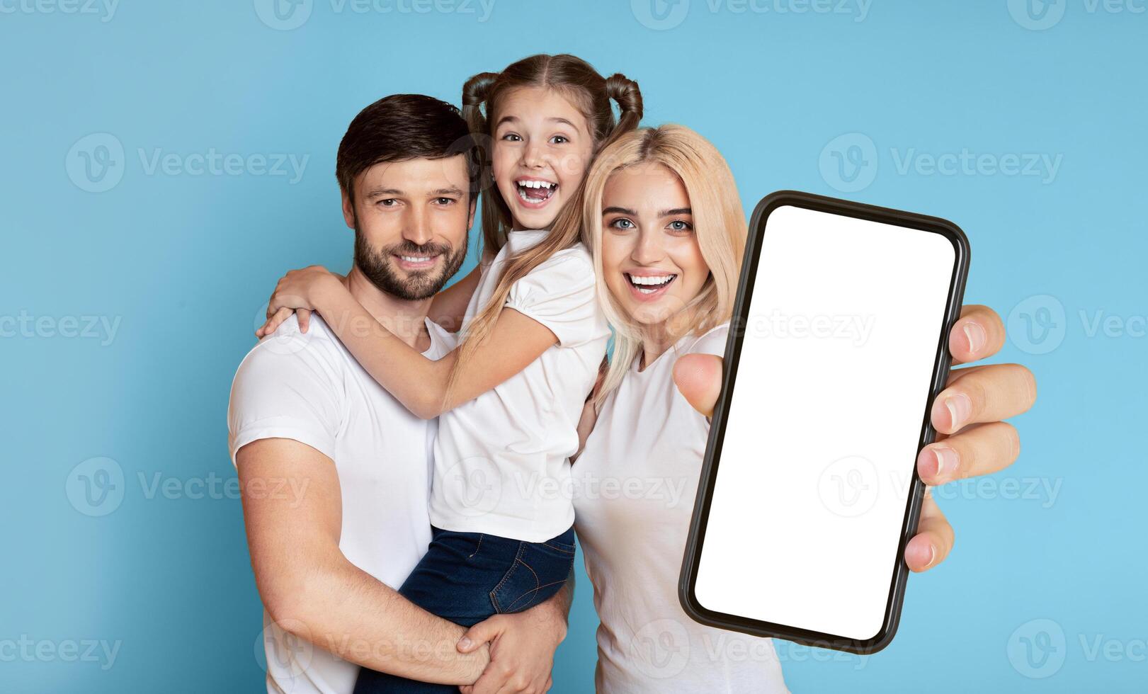 A happy family, consisting of a father, mother, and daughter, are standing in front of a blue background. The father is holding the daughter in his arms, and the mother is holding a smartphone photo
