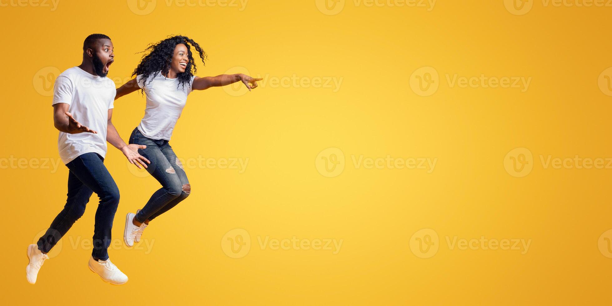 Surprised Black Man Is Looking At Free Space, afro woman is jumping and pointing at empty space, yellow background, panorama photo