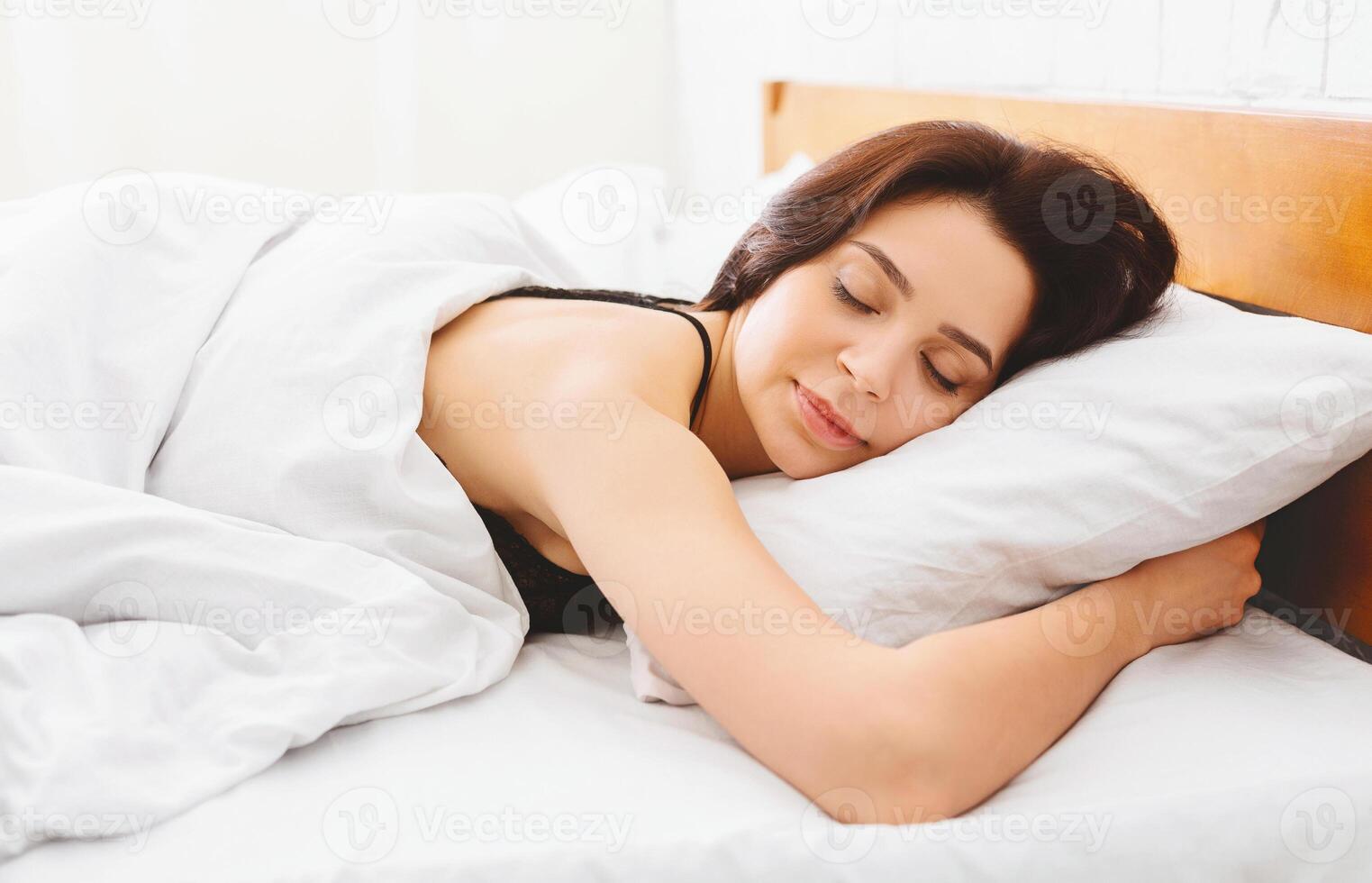 A woman is peacefully asleep in a bed with crisp white sheets covering her. The room is softly lit, creating a cozy and restful atmosphere. photo