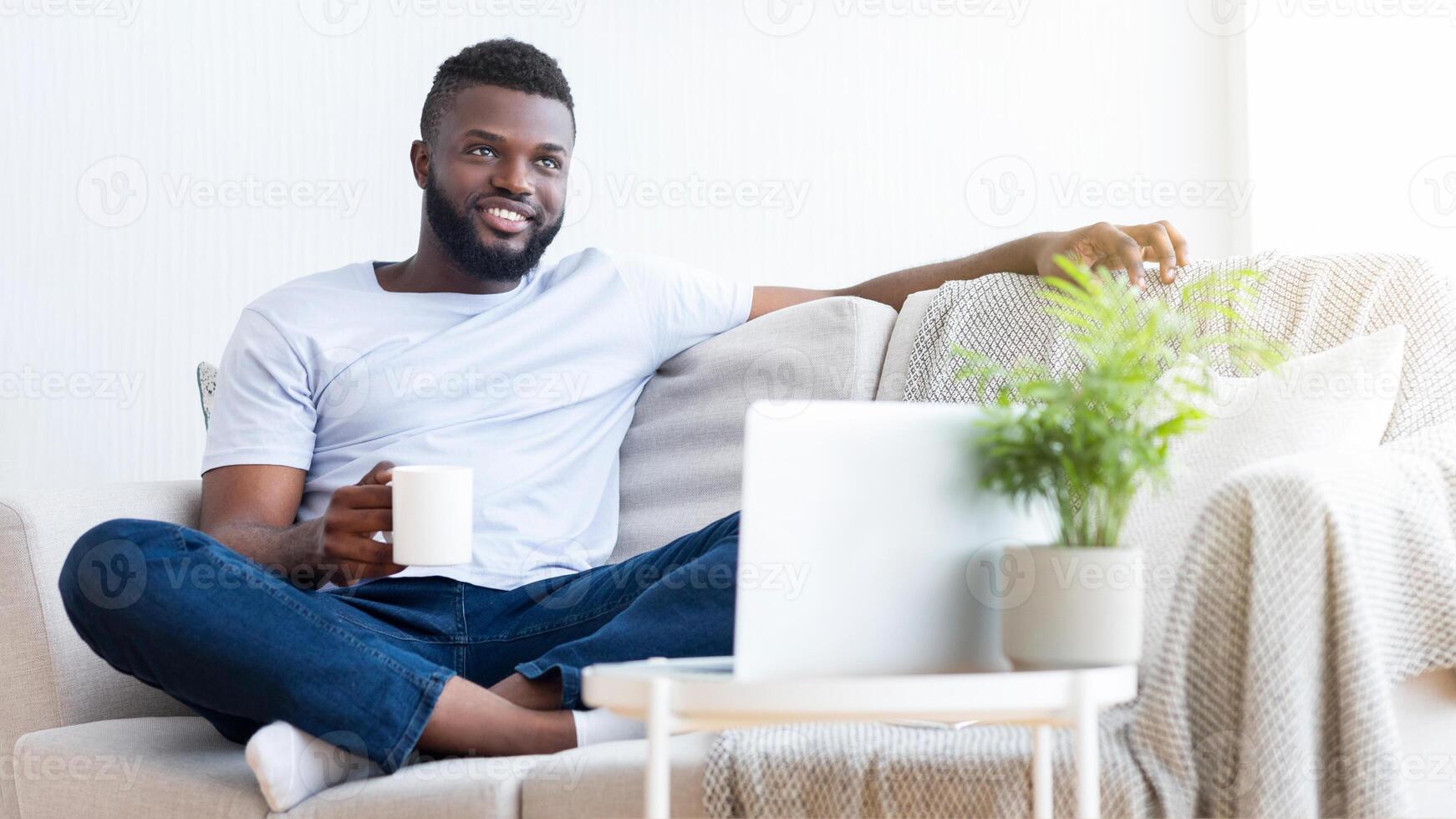 A young black man in a white t-shirt and jeans relaxes on a couch in his living room, holding a white mug of coffee and gazing out the window, copy space photo