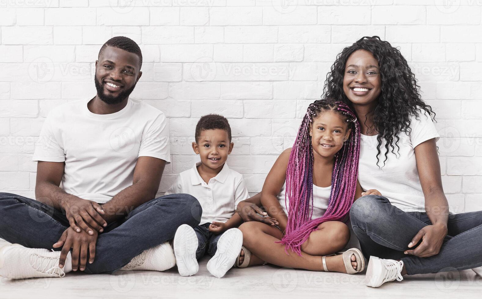 Family love and care. Smiling black parents with their two little kids sitting on floor together and looking at camera over white wall background photo