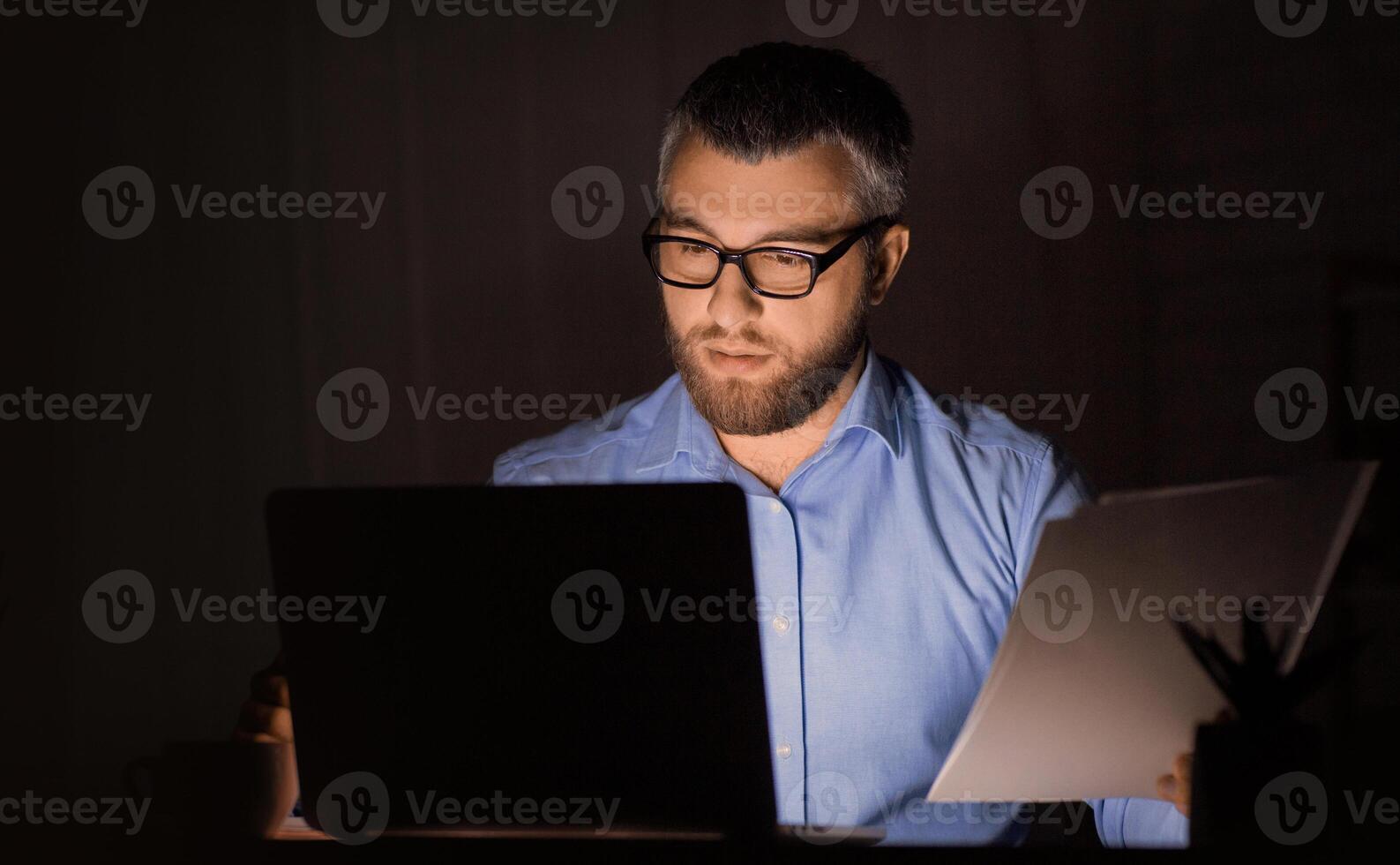 A man wearing glasses and a blue shirt is working late at night in a dimly lit office. He is concentrating on both his laptop and a document he is holding in his hand. photo