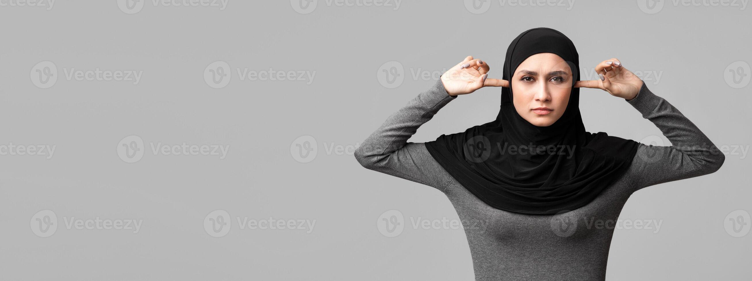 Hear no evil. Arabic girl in hijab closing ears with fingers and looking at camera with angry face expression, gray background with free space photo