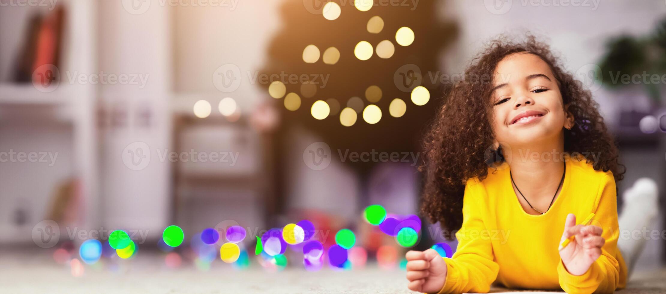 Xmas fun. Cheerful african little girl drawing on floor near decorated Christmas tree at home, free space photo