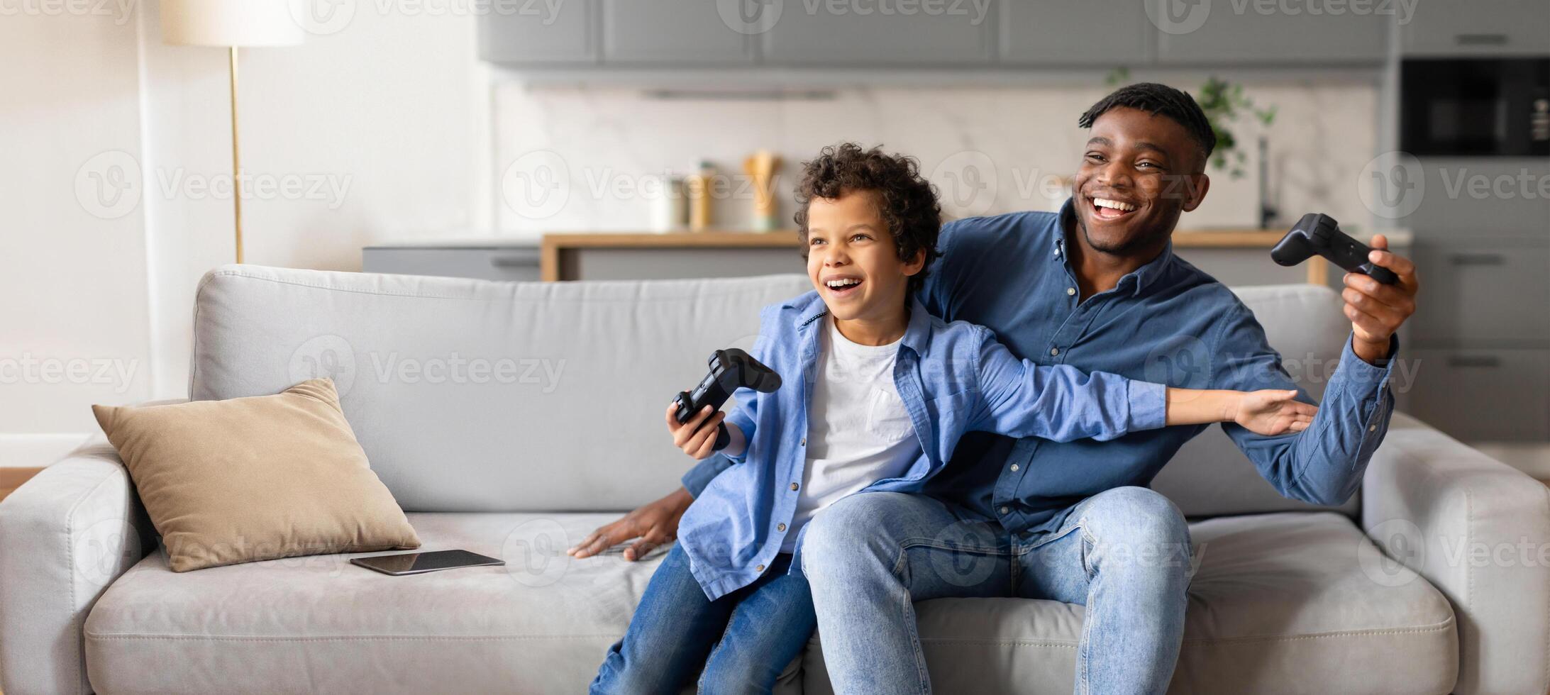 African American father and son share laughter while playing games together on a comfortable sofa. The bright living room is filled with warmth and joy during their bonding time. photo