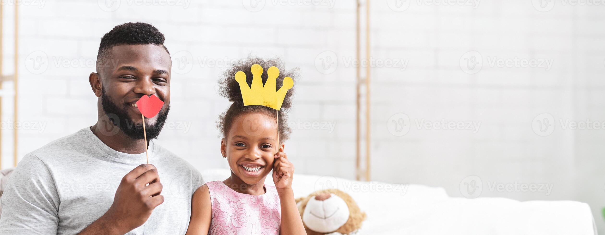 A joyful black father and his daughter are sharing a lighthearted moment, both smiling and posing for fun with playful props. The father holds a red heart-shaped object, while the girl wears a crown photo