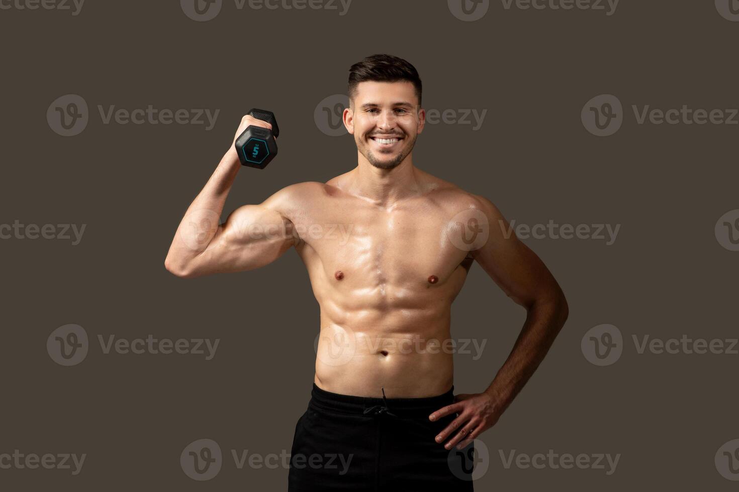 A man is standing and posing while holding a dumbbell in his hand. He is showcasing his muscles and strength, with a focused expression on his face. photo