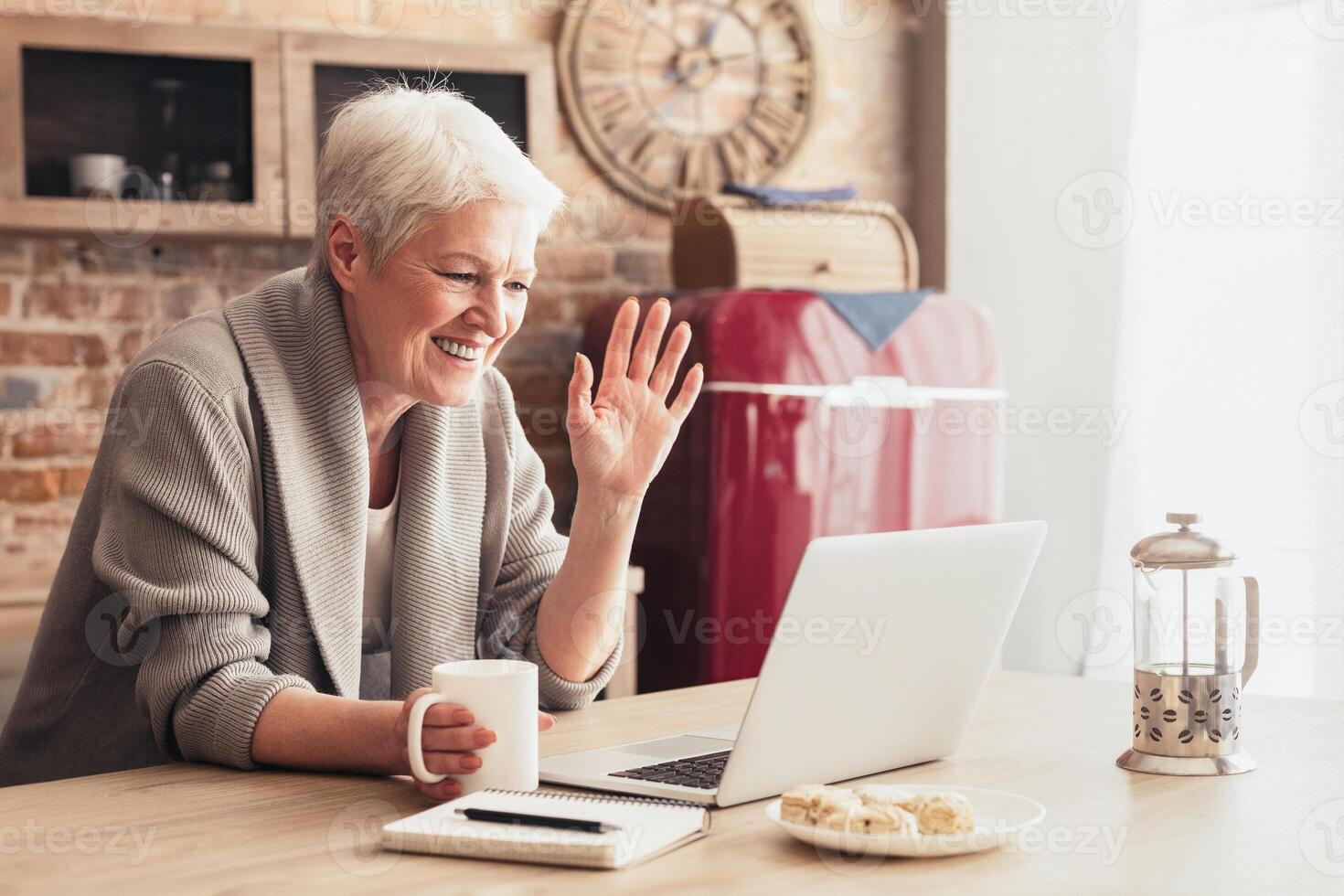 A senior woman sits at a kitchen table with a laptop in front of her. She is holding a cup of coffee in her left hand and waving with her right hand, indicating she is chatting with someone. photo