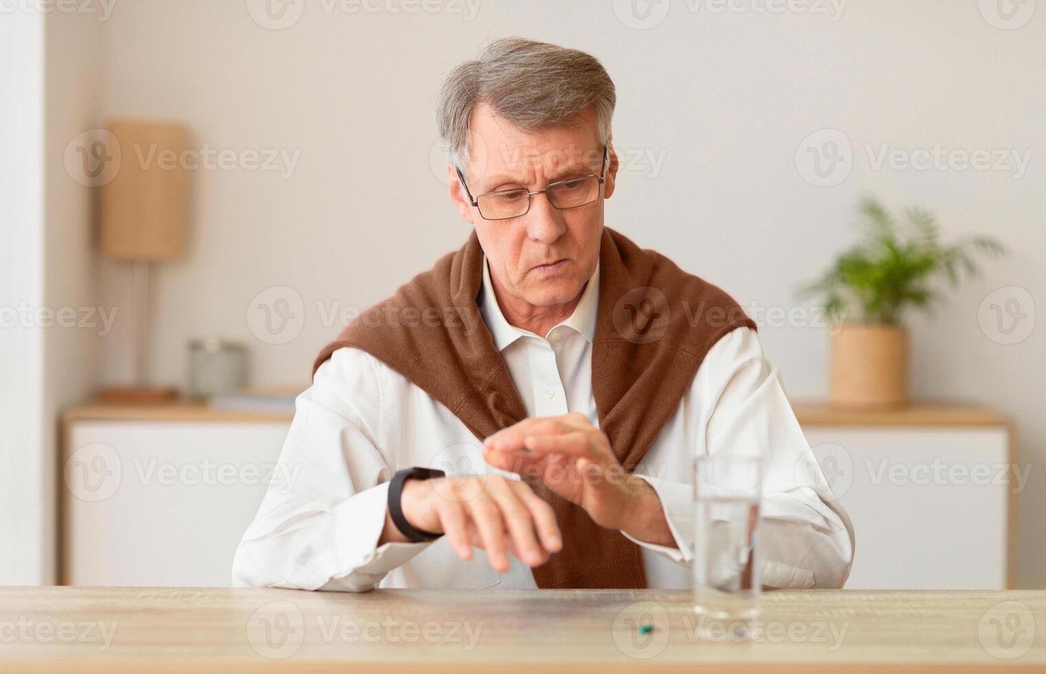 Elderly Gentleman Checking Time With Smartwatch Sitting At Table With A Pill And Glass Of Water At Home photo