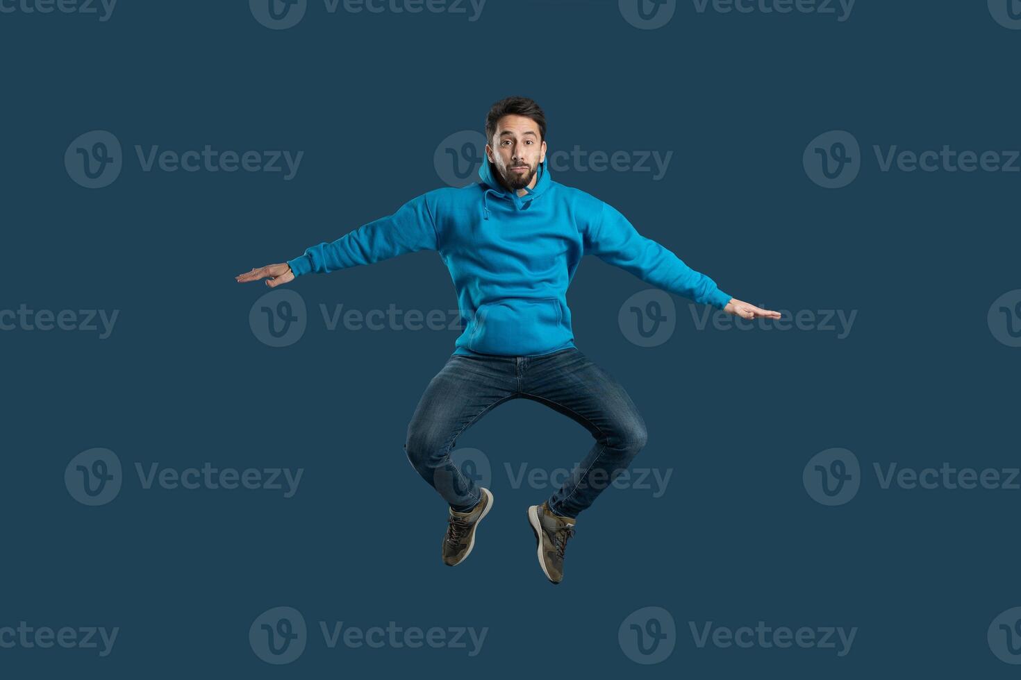 A young man demonstrates his enthusiasm by leaping off the ground in a bright blue hoodie, showcasing his playful spirit and joy. The solid dark background emphasizes his energetic pose photo