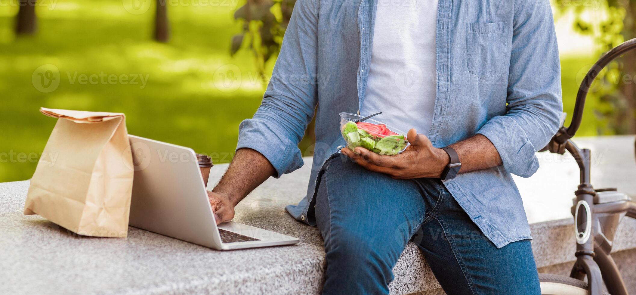 Business Lunch On Fresh Air. Unrecognizable African Man Eating Salad And Using Laptop, Resting On Bench In Park, Panorama, Cropped Image photo
