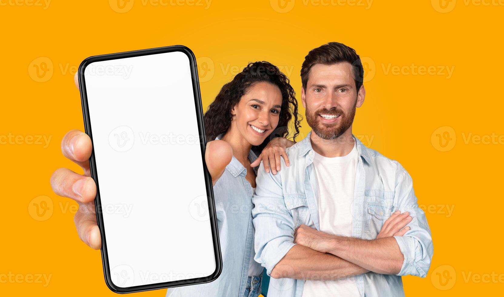 A couple stands in front of a bright yellow background. The woman has her arm around the man's shoulder and they both smile at the camera. The woman holds a smartphone with a blank screen photo