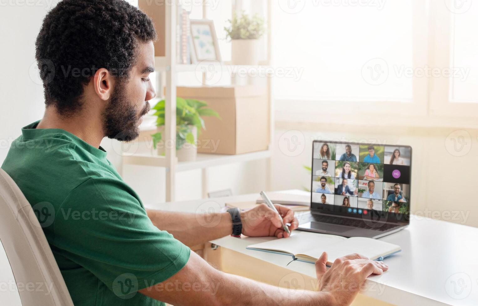A man sitting at his desk in a home office during the daytime, actively participating in a virtual business meeting on his laptop while taking notes in a notebook. photo