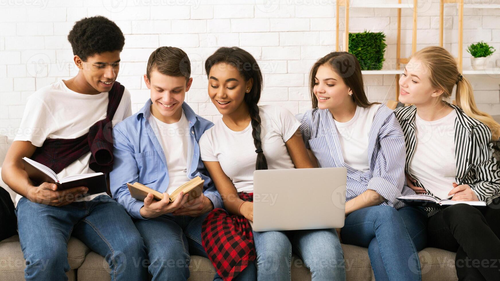 Five multiethnic teenagers are sitting closely together, sharing a casual study session indoors. Two of them are reading books, one is typing on a laptop, and the others appear to be discussing photo