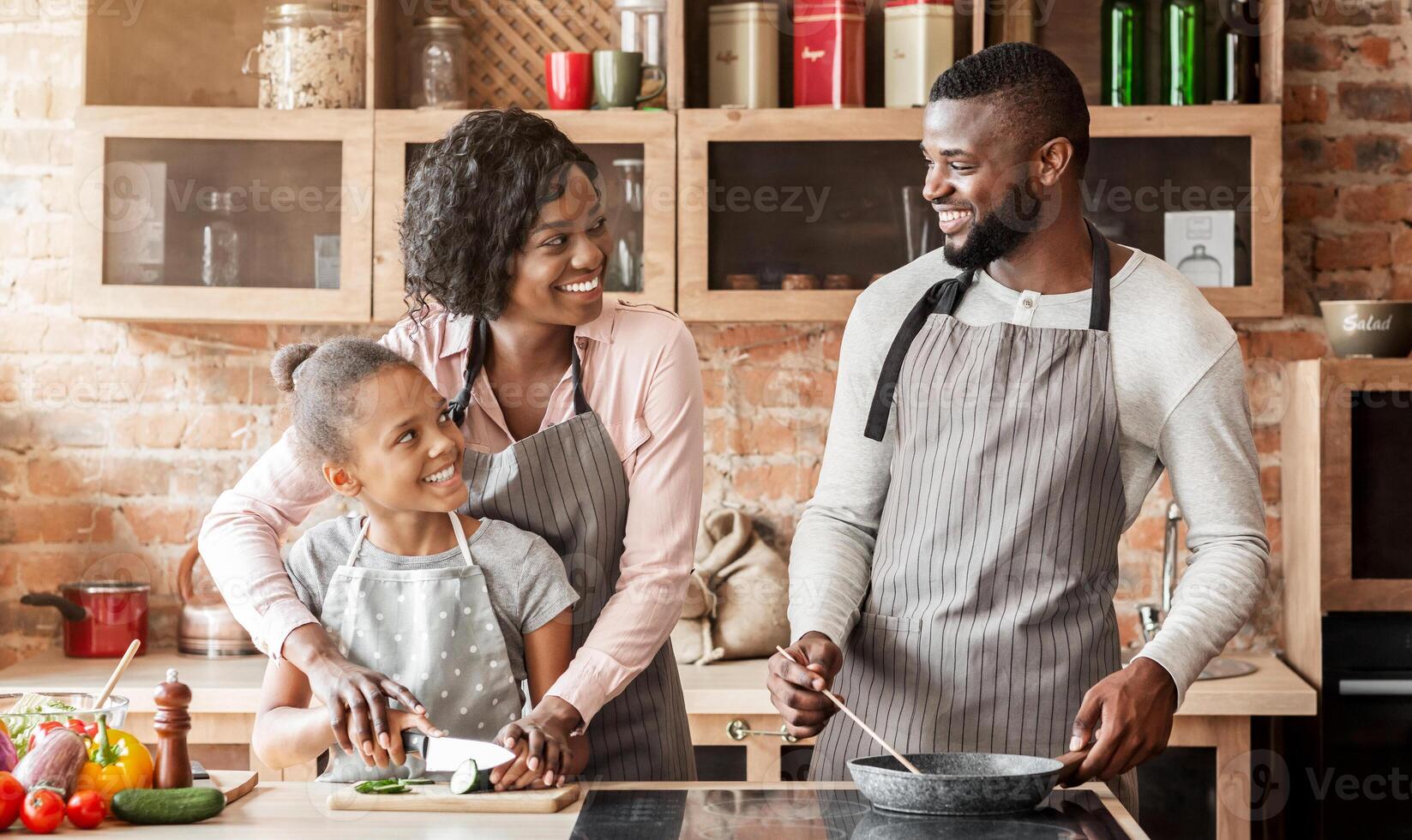 Smiling african mother teaching little daughter cutting vegetables, cheerful father frying food, kitchen interior photo