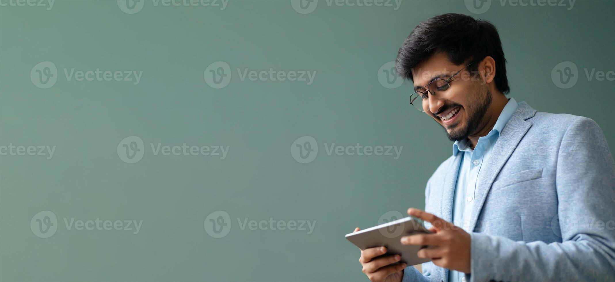 Indian man is engaged with a tablet, smiling warmly in a contemporary indoor space. He wears a light suit and glasses, creating a relaxed and professional atmosphere. photo