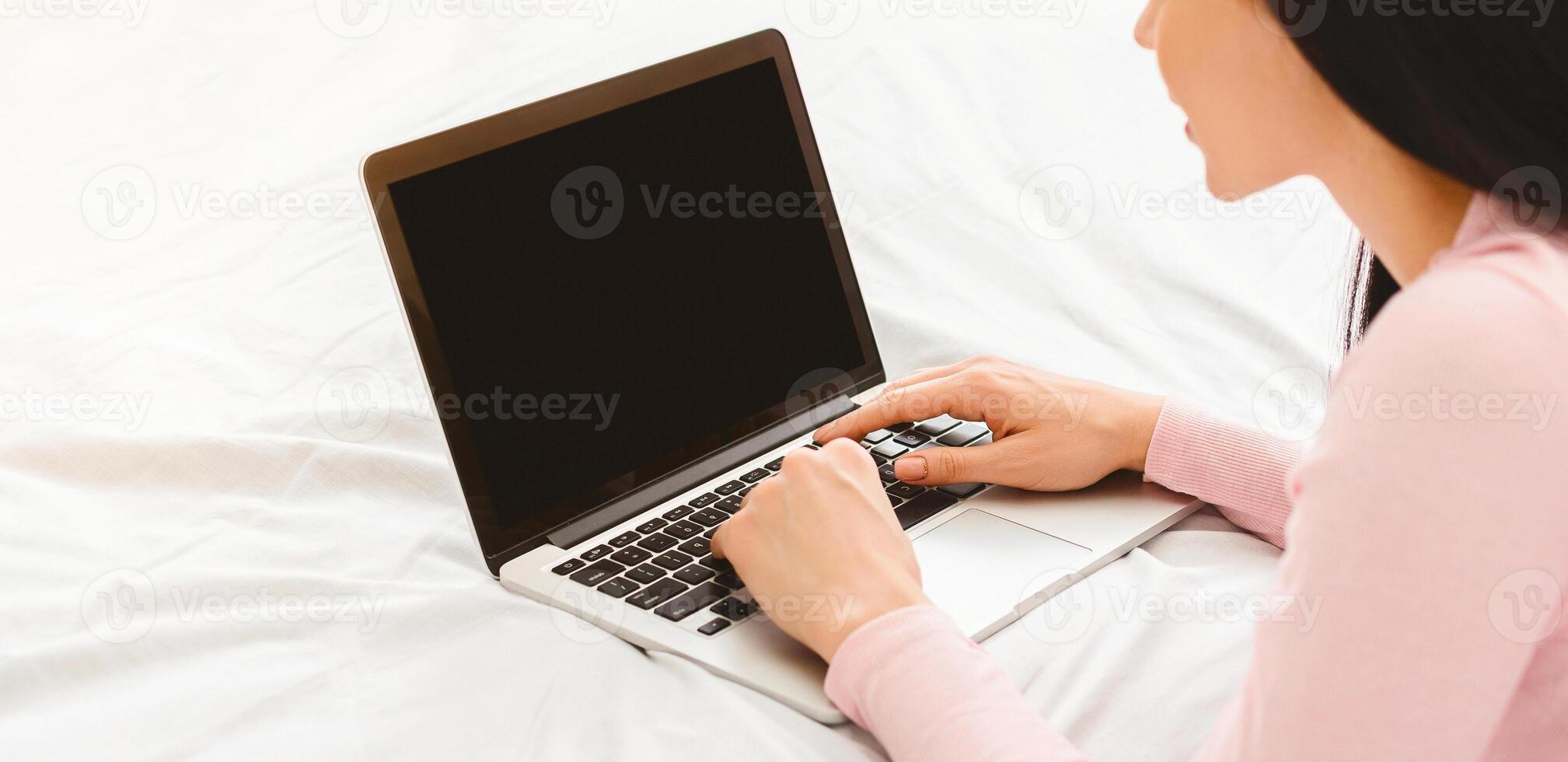 A woman is seated on a bed, attentively using a laptop computer with blank screen in front of her. She appears focused on her screen, typing and clicking, cropped photo