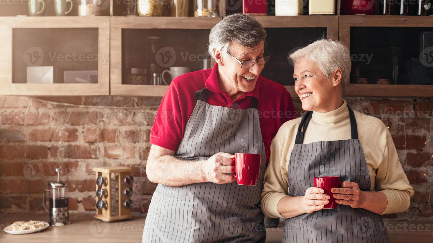 A senior couple wearing aprons stands together in a kitchen, each holding a red mug of coffee. They are smiling and looking at each other, enjoying a moment of connection and togetherness. photo