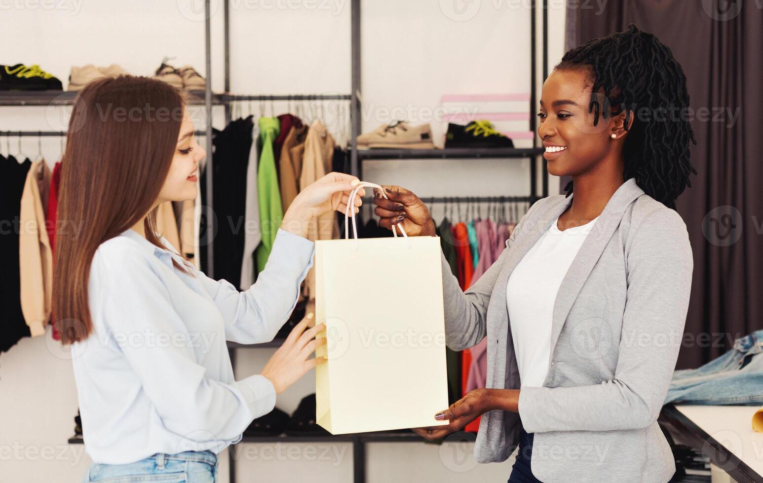 Two multiracial women are browsing through racks of clothes in a busy clothing store. They are holding clothing items and discussing their choices photo