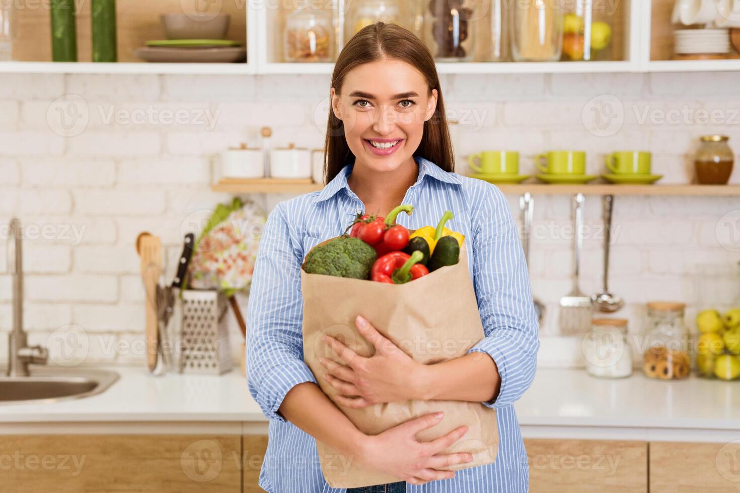 A woman stands in her kitchen, smiling, and holding a brown paper bag filled with fresh produce. photo