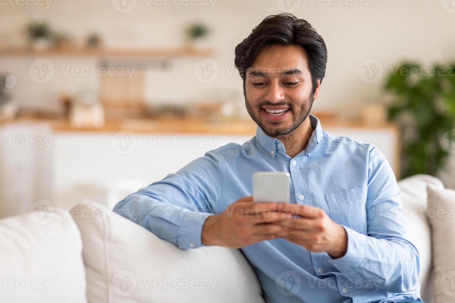 Arab man sits on a white couch and looks at his phone with a smile on his face. The man is wearing a blue button-down shirt. The background is out of focus and shows a kitchen area. photo