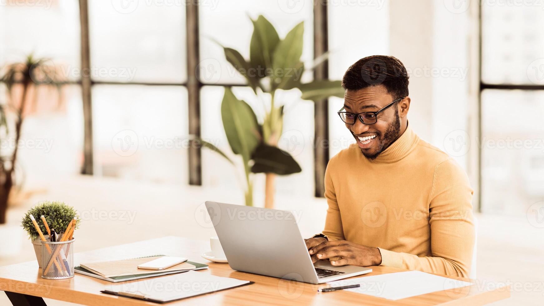 Excited Guy. Happy black man in glasses working on laptop to get all his business done early in the morning, panorama photo